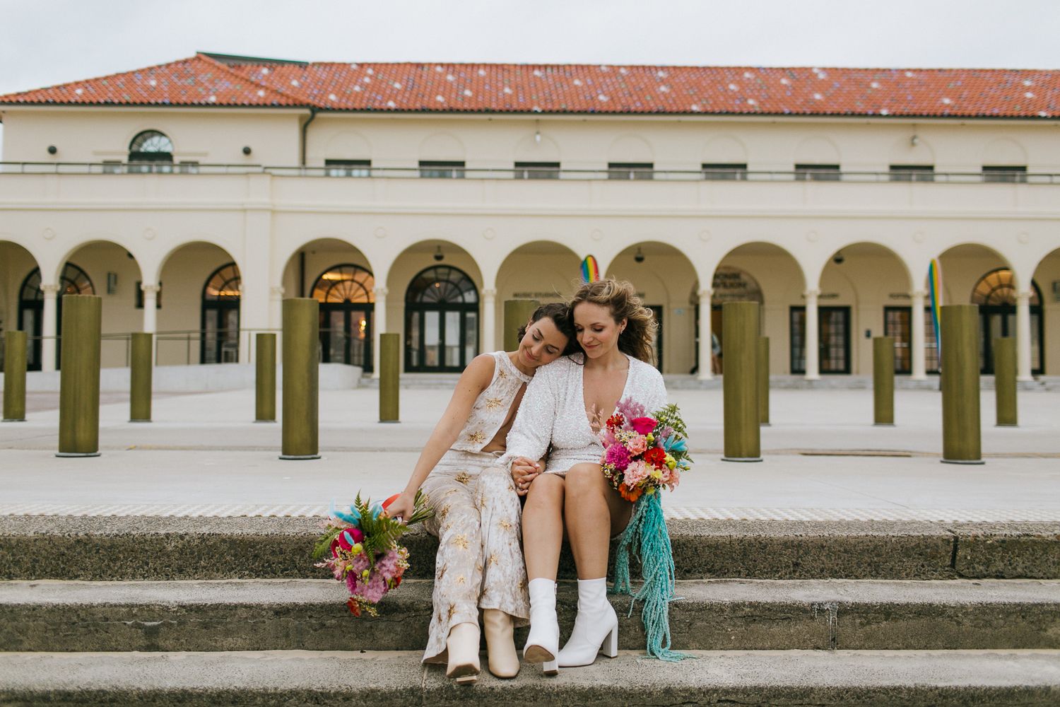 Two brides sitting on steps holding vibrant bouquets at elegant venue.