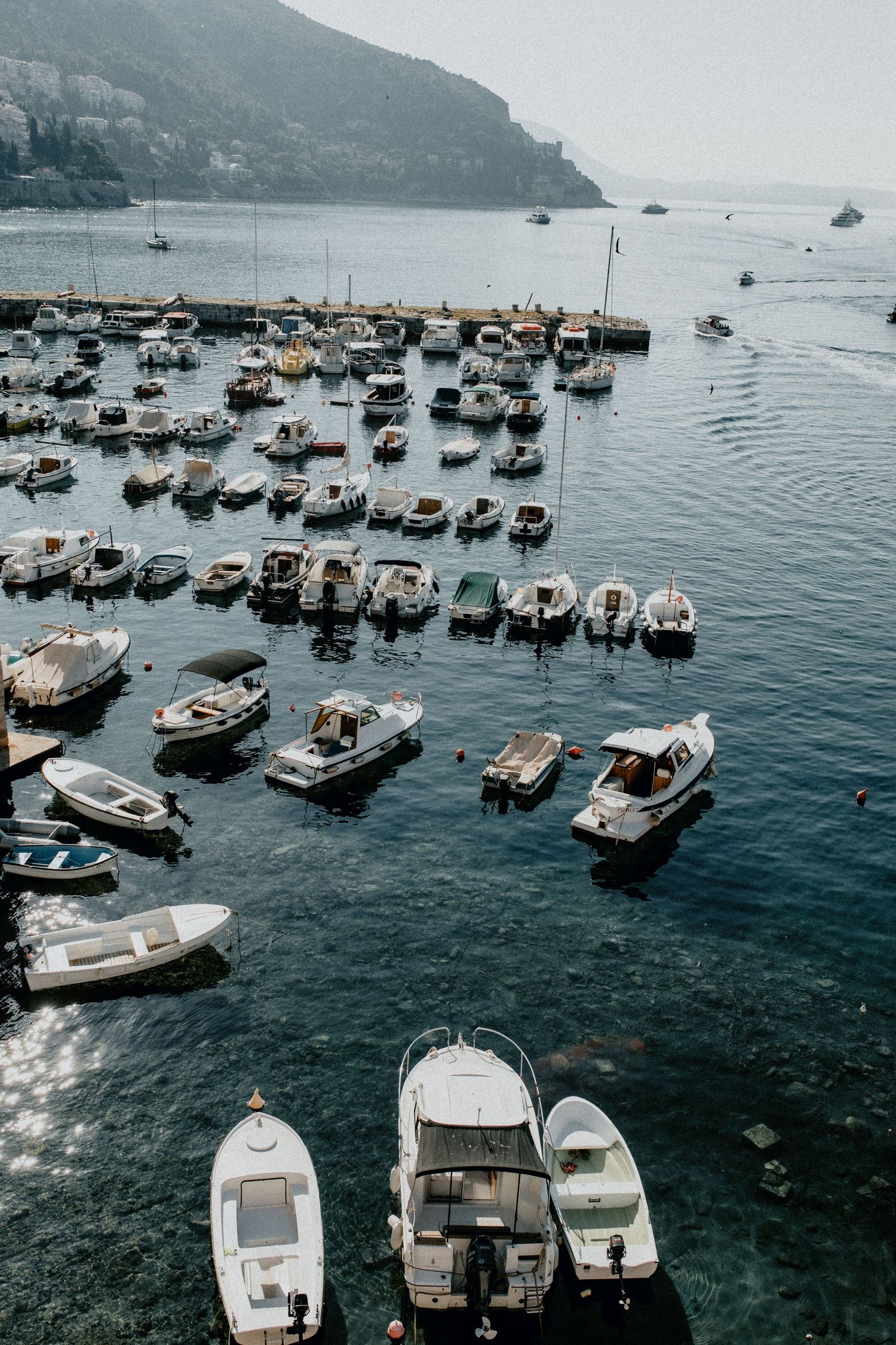 Aerial view of small boats and yachts moored in a Mediterranean harbor surrounded by mountains.