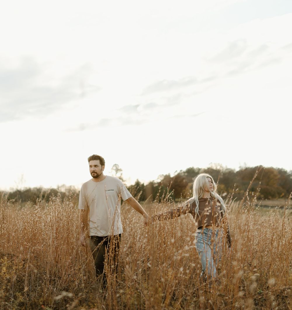 A couple holds hands while standing in a golden field during sunset.