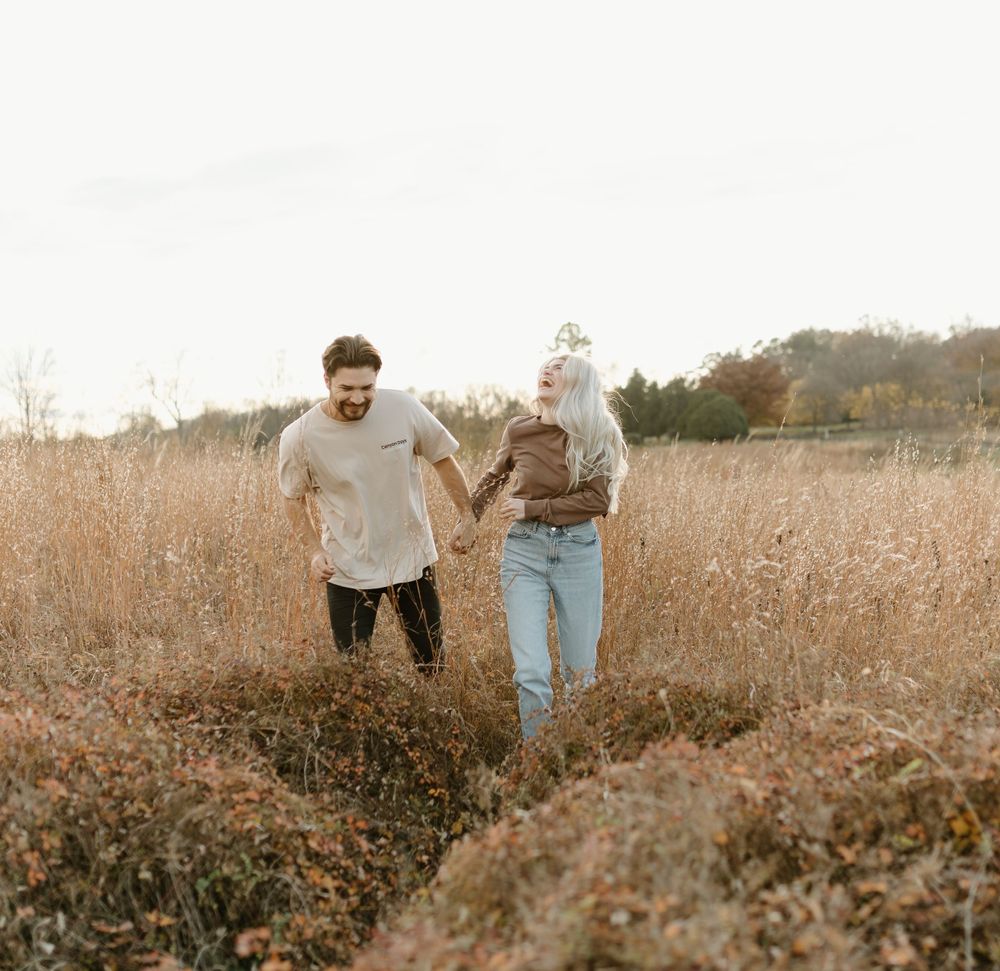 A couple runs playfully through a golden field during autumn.
