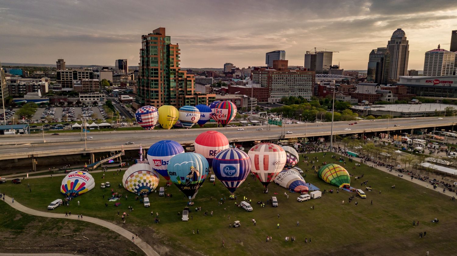 Hot air balloons gathered at Louisville waterfront park with city skyline in background at sunset.
