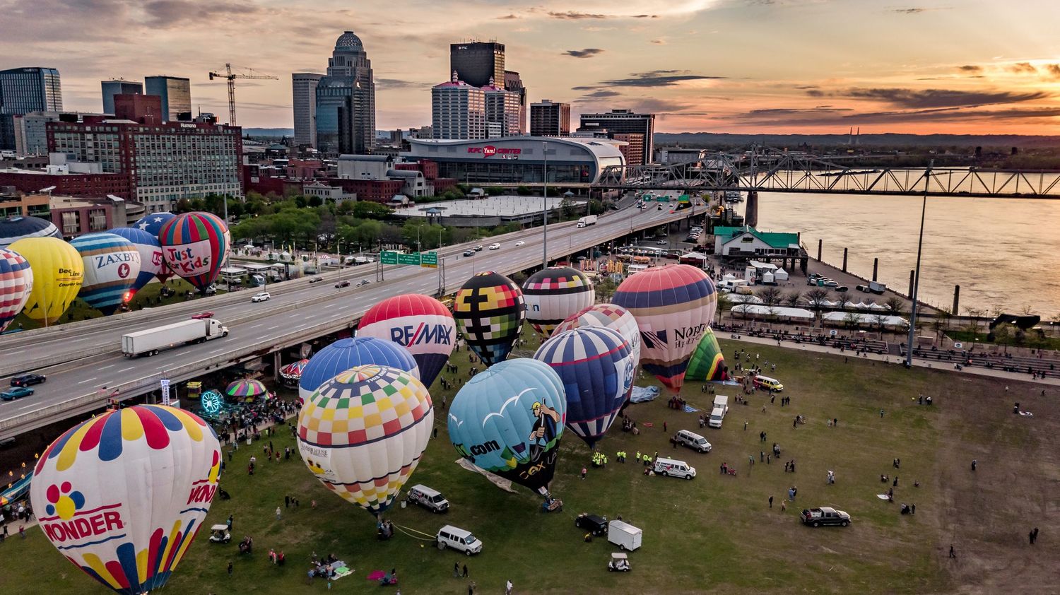 Wide aerial view of Louisville waterfront festival with colorful hot air balloons and city buildings.