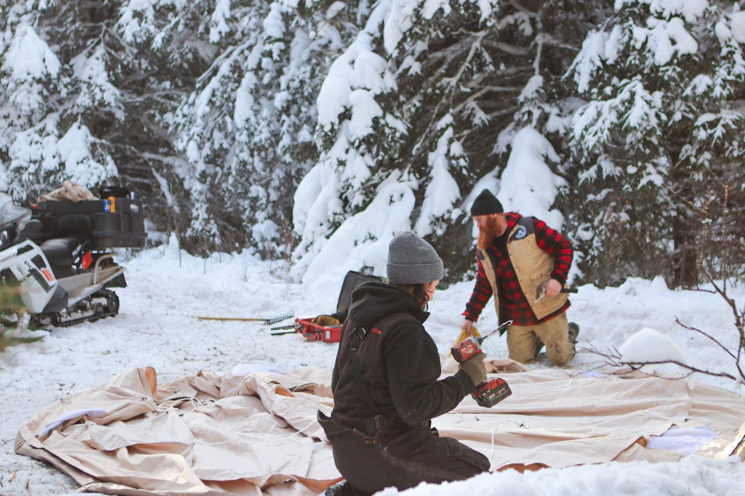 Setting up winter camping equipment on a tarp in snowy forest conditions.