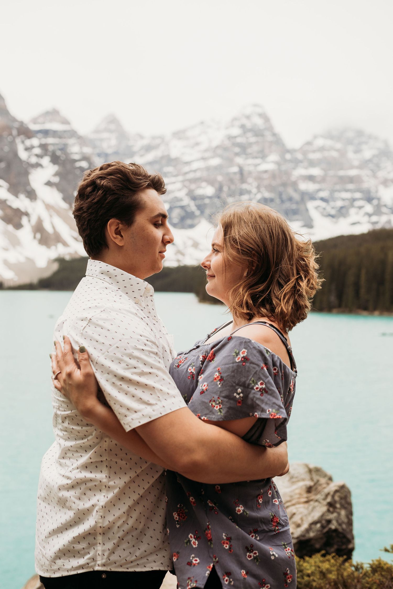 A couple embraces beside turquoise waters of Moraine Lake with snow-covered peaks in the background.