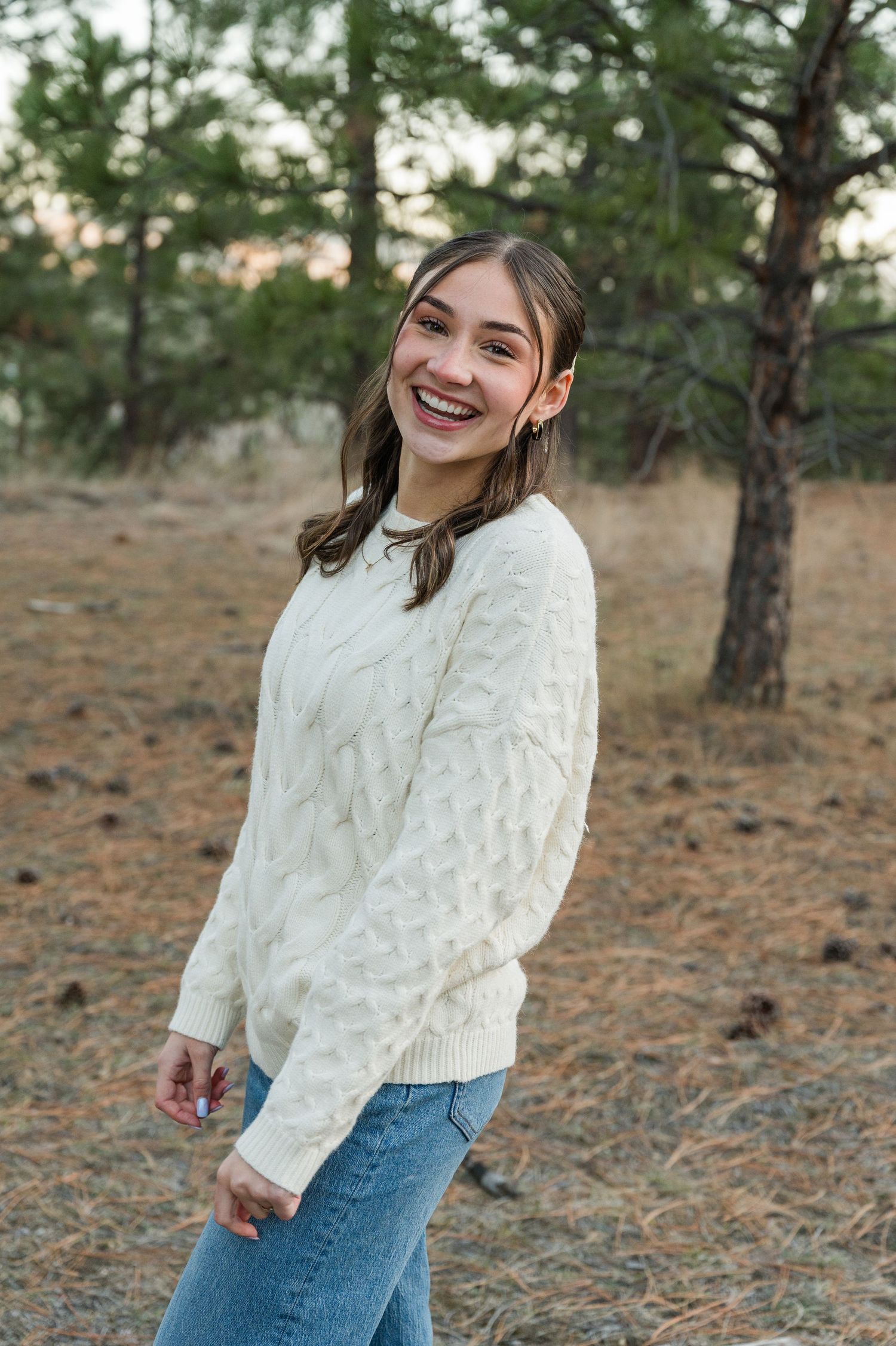 Person wearing a cozy white cable knit sweater smiles joyfully in a rustic outdoor setting with pine trees.