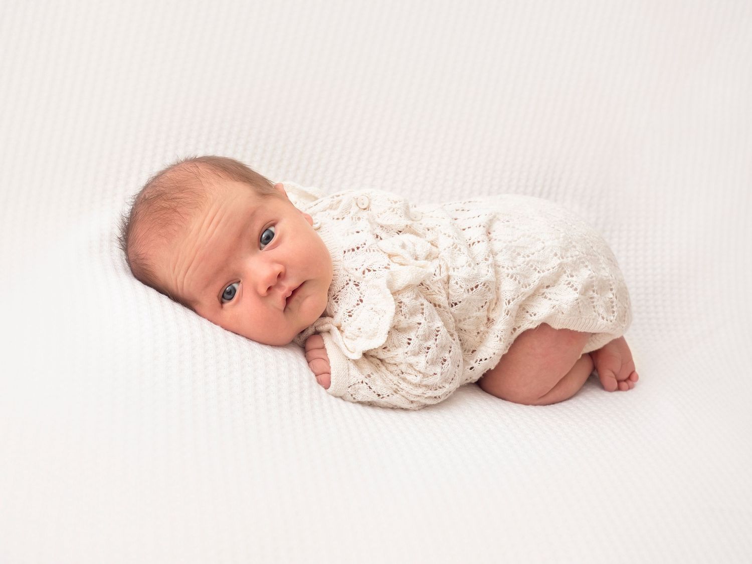 A newborn baby girl in a cream outfit lays posed on her tummy, awake and looking at the photographer.