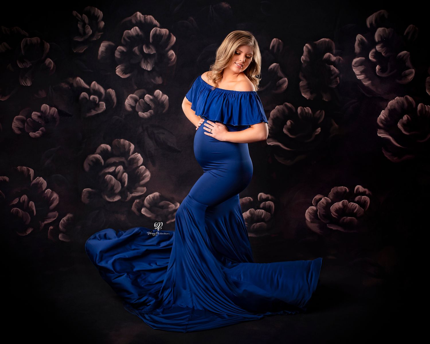 Blonde pregnant woman wearing blue maternity dress poses with hands on belly, fabric flowing; floral backdrop; maternity