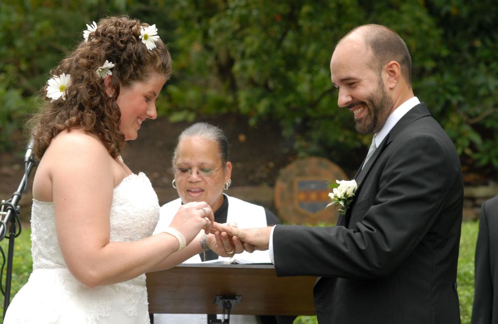 A couple exchanges rings during an outdoor wedding ceremony with an officiant presiding at a wooden podium.