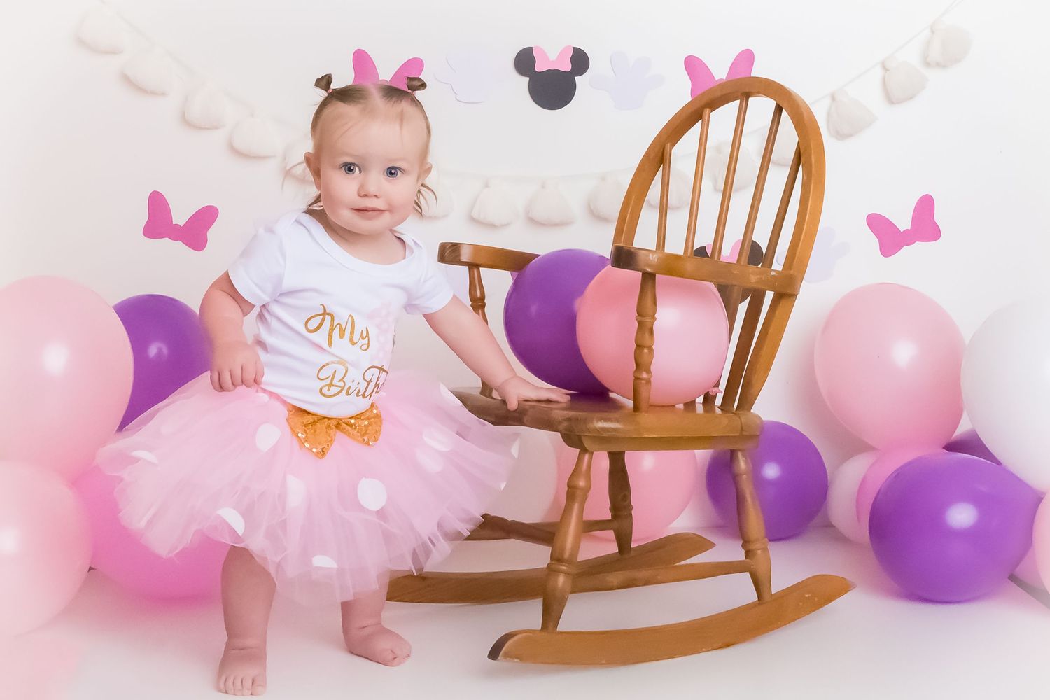 Pink and purple balloon decorated scene with tutu outfit and wooden rocking chair for special celebration.