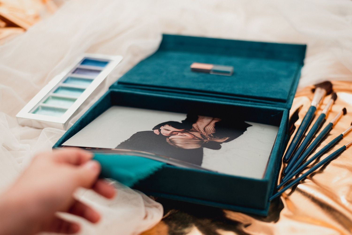 Hands opening a teal velvet box containing photographs and art supplies.