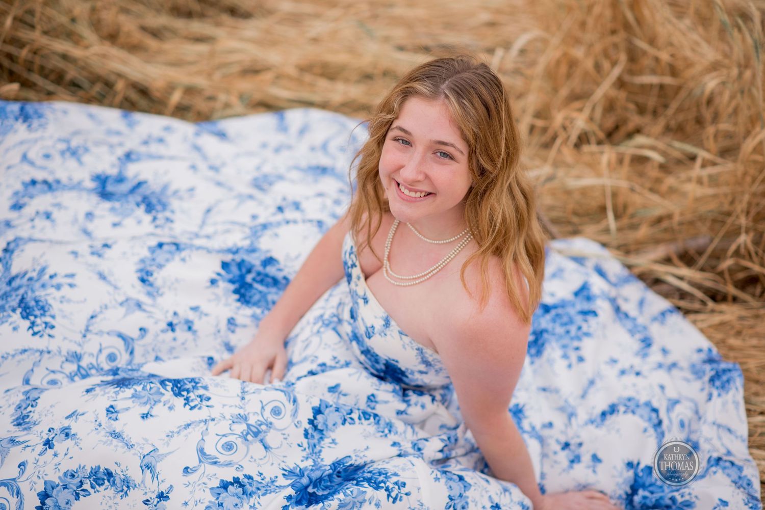 Joyful portrait in a blue and white floral gown sitting amidst golden hay bales.