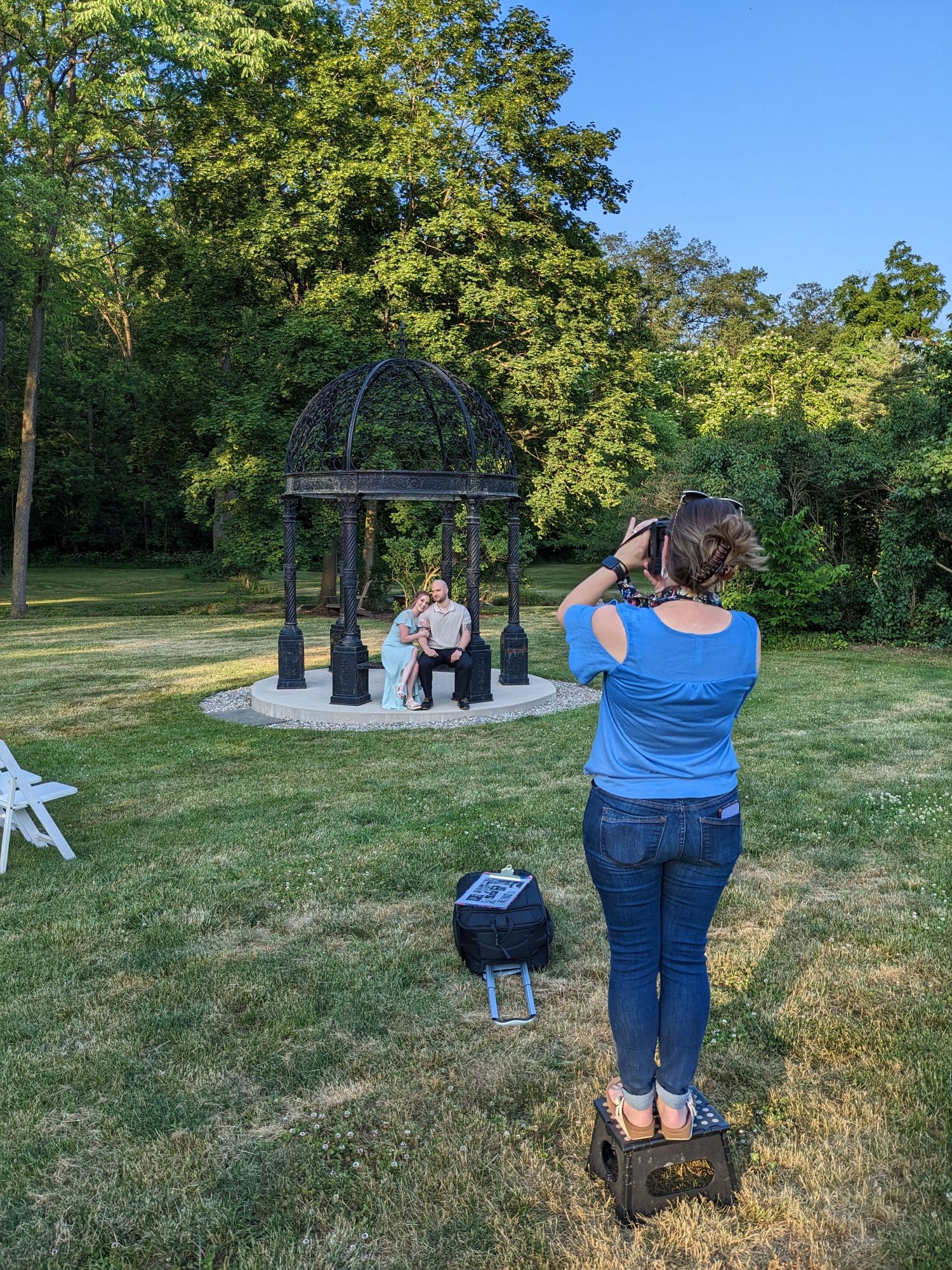 A photographer captures a wedding ceremony under a gazebo in a sunny garden setting.