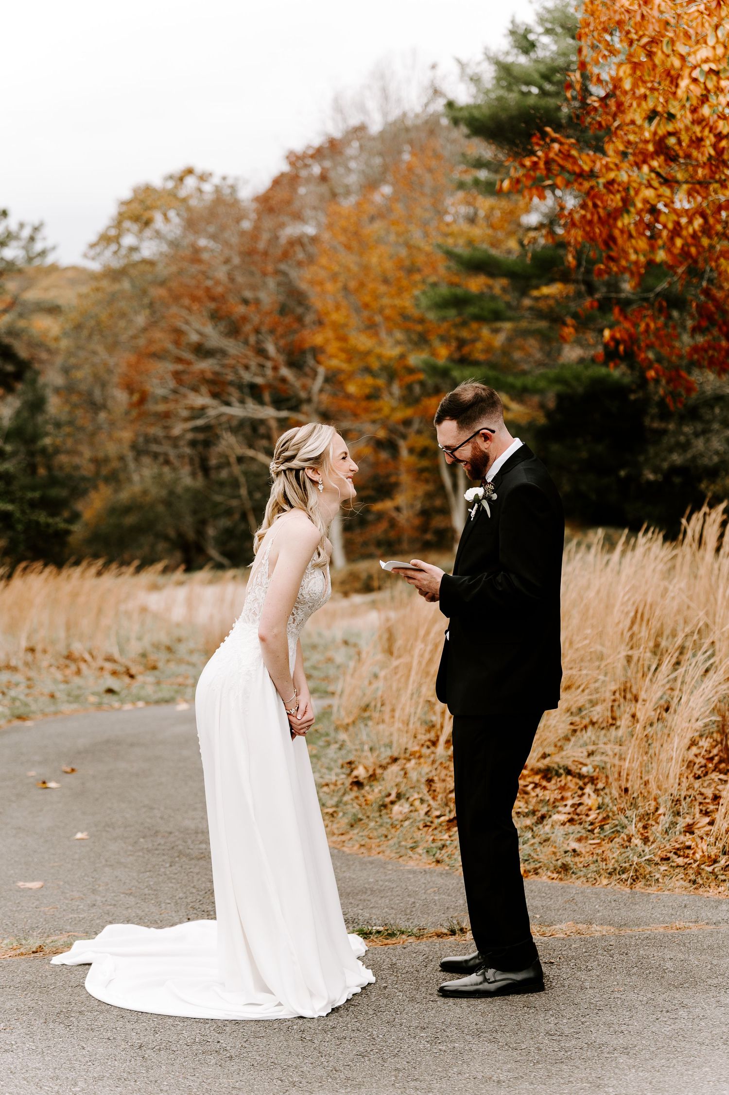 A wedding couple stands on a path surrounded by fall colors as they read personal letters to each other.