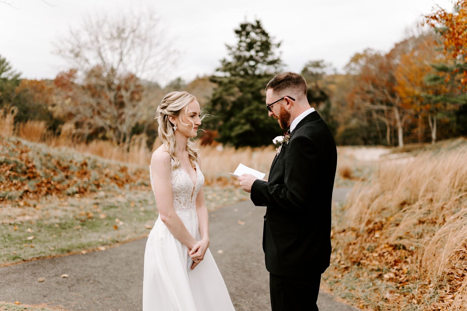 A couple stands together on a path surrounded by autumn trees during their first look.