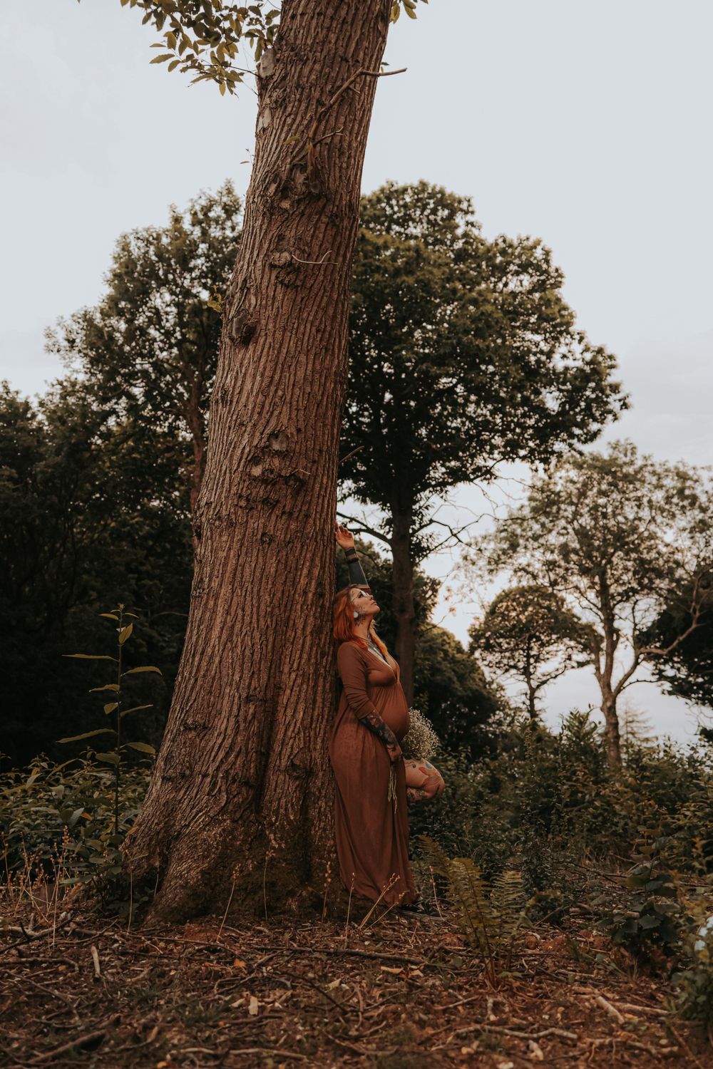 A dramatic figure in a flowing dress poses beside a large tree trunk in moody lighting.