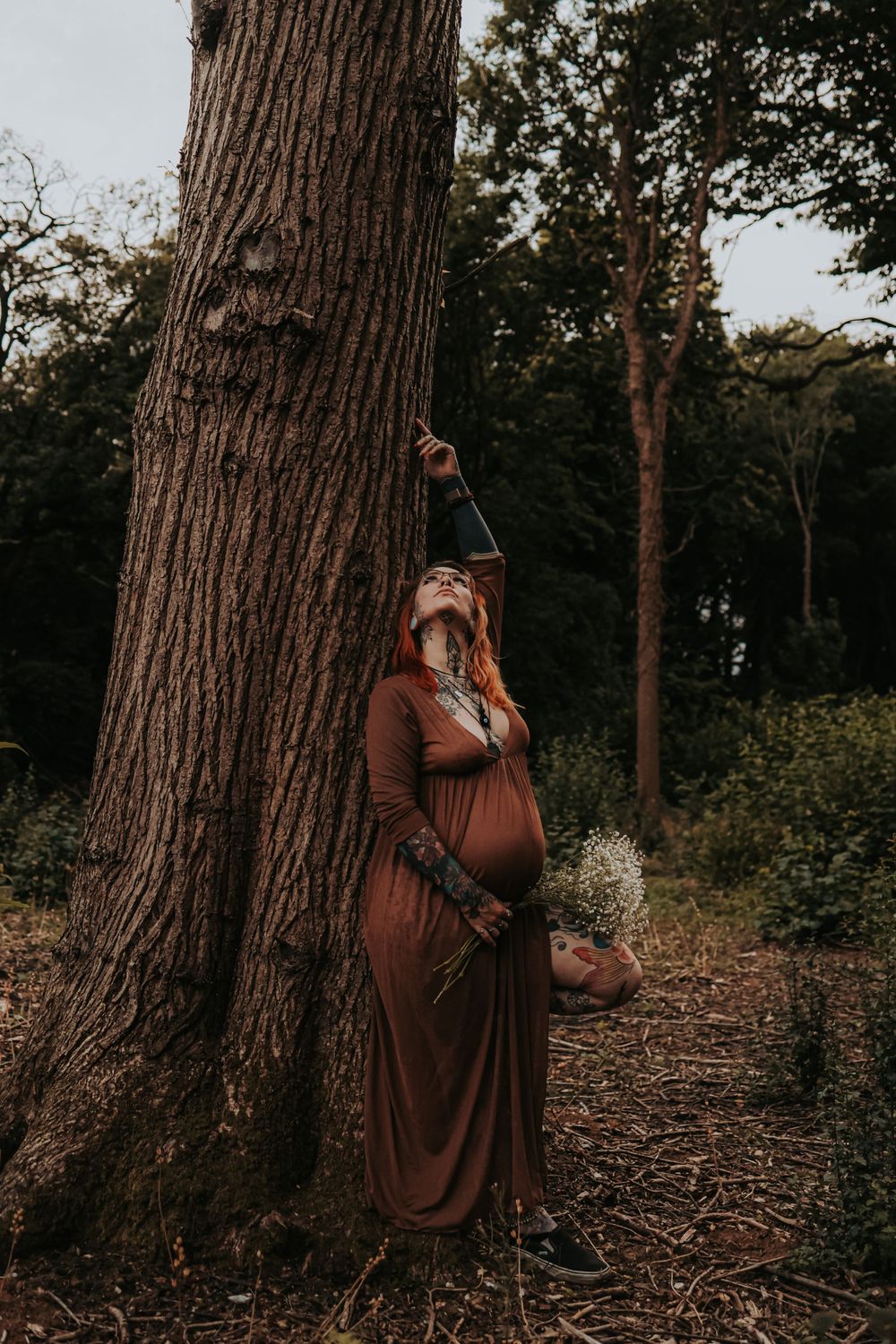 A maternity photo featuring a person in a brown dress against a large tree in the woods.