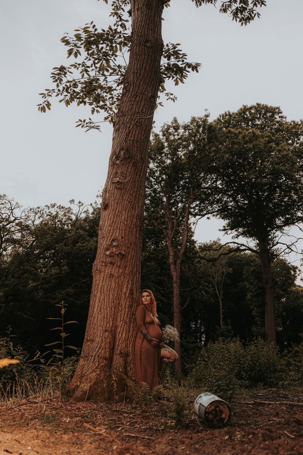 A figure in a rust-colored dress stands beside a massive tree trunk in a wooded setting.