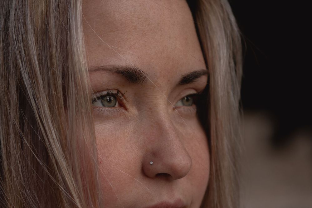 Close-up portrait showing blonde hair and a nose piercing against a dark background.