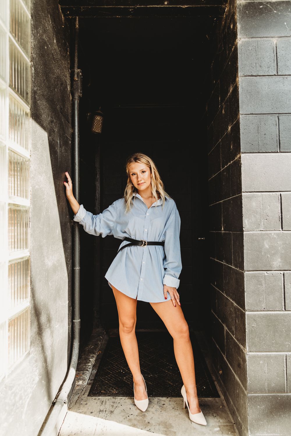 Dramatic fashion poses against dark industrial doorway wearing oversized blue shirt.