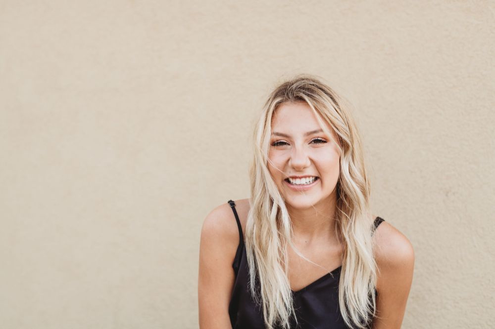 Woman in black tank top smiling against beige wall background.