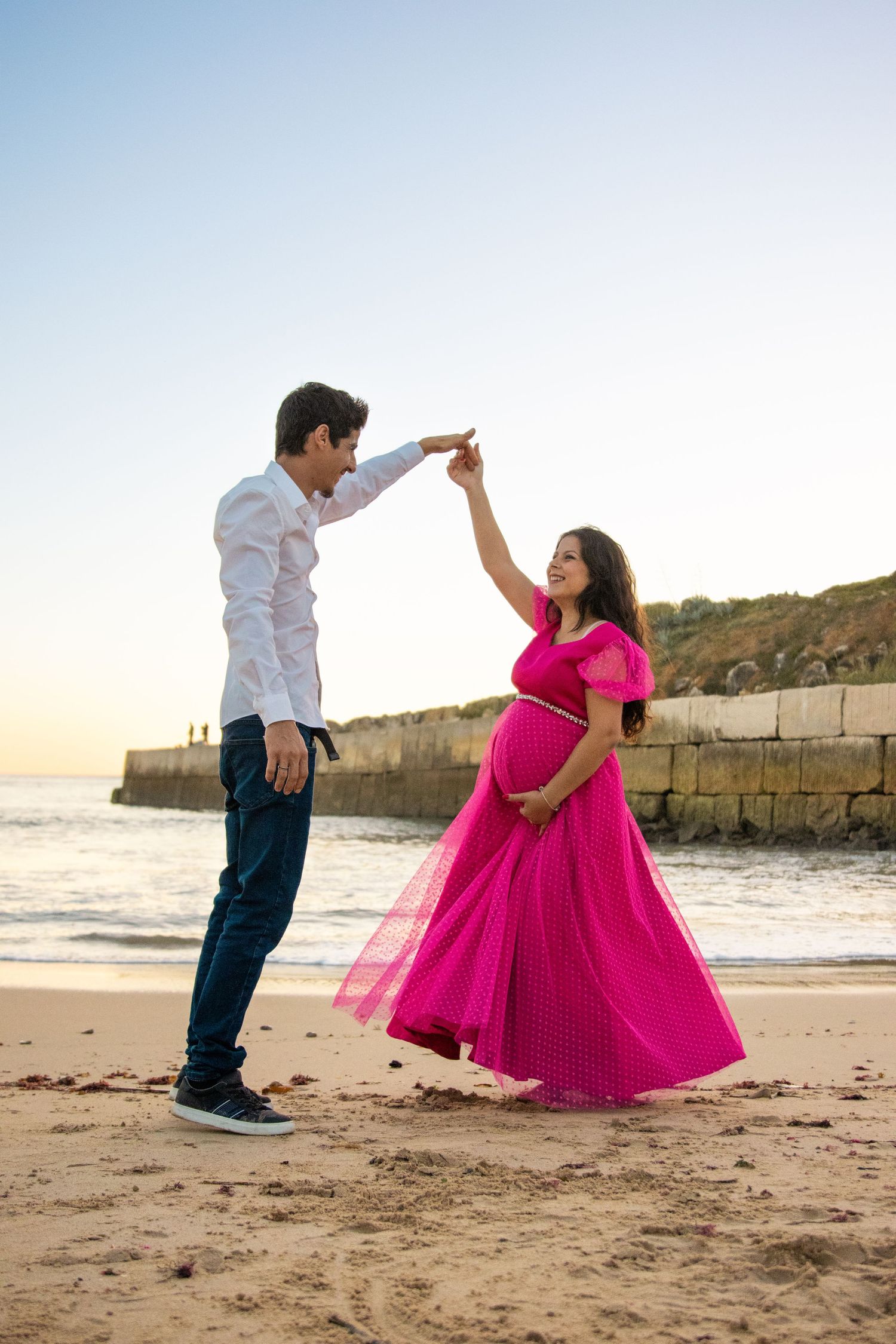 Couple dances playfully on a beach at sunset with pink flowing dress creating movement.