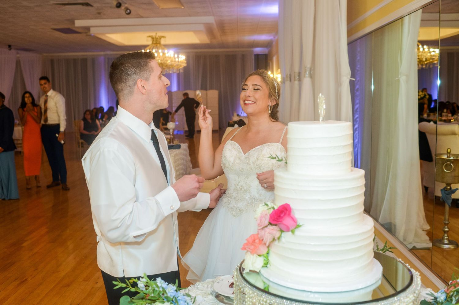 Reception moment with wedding cake and couple sharing a slice in an elegant ballroom.