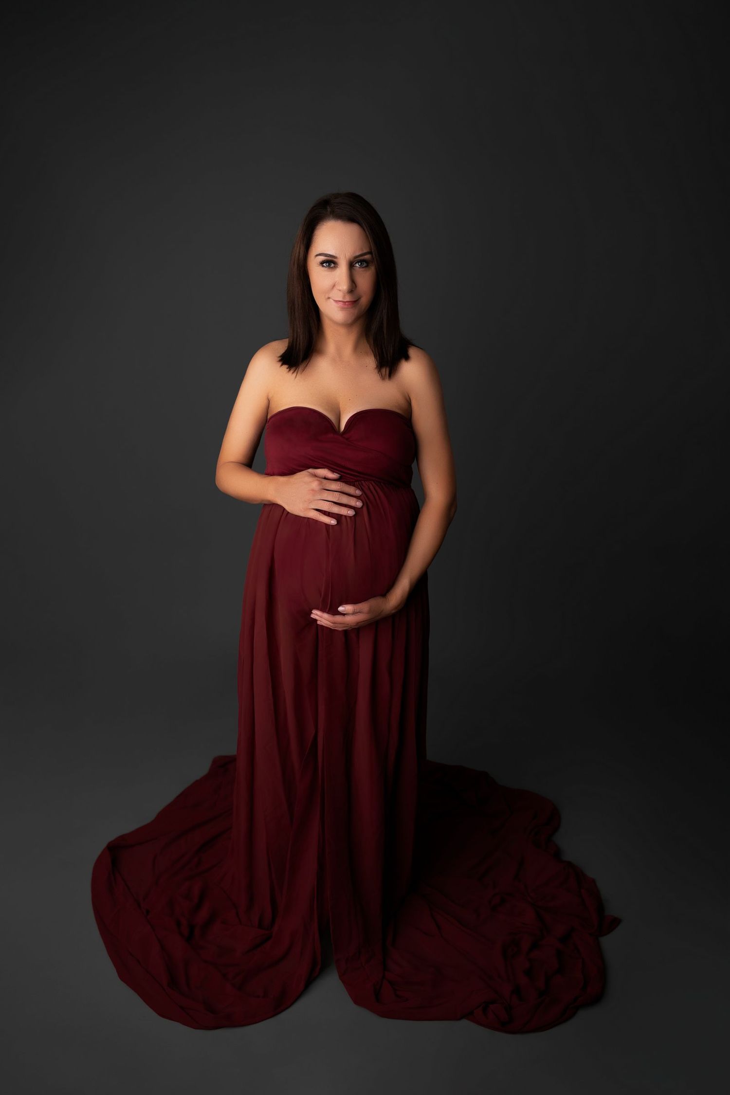 A burgundy maternity gown flows elegantly against a dark background in this dramatic studio portrait.