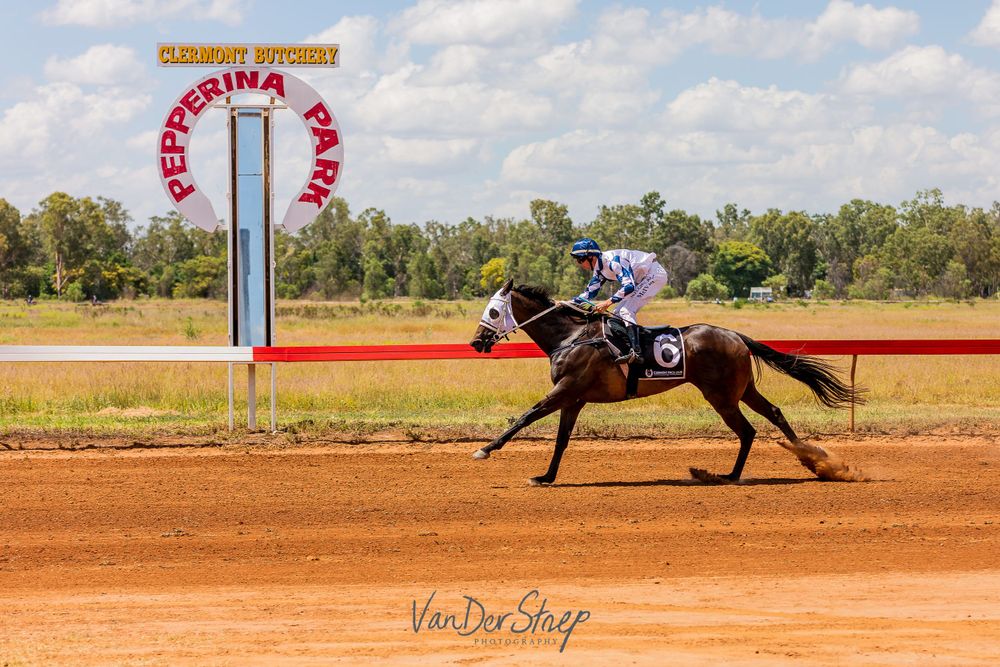 Racing horse and jockey sprinting past finish line arch on dirt track.