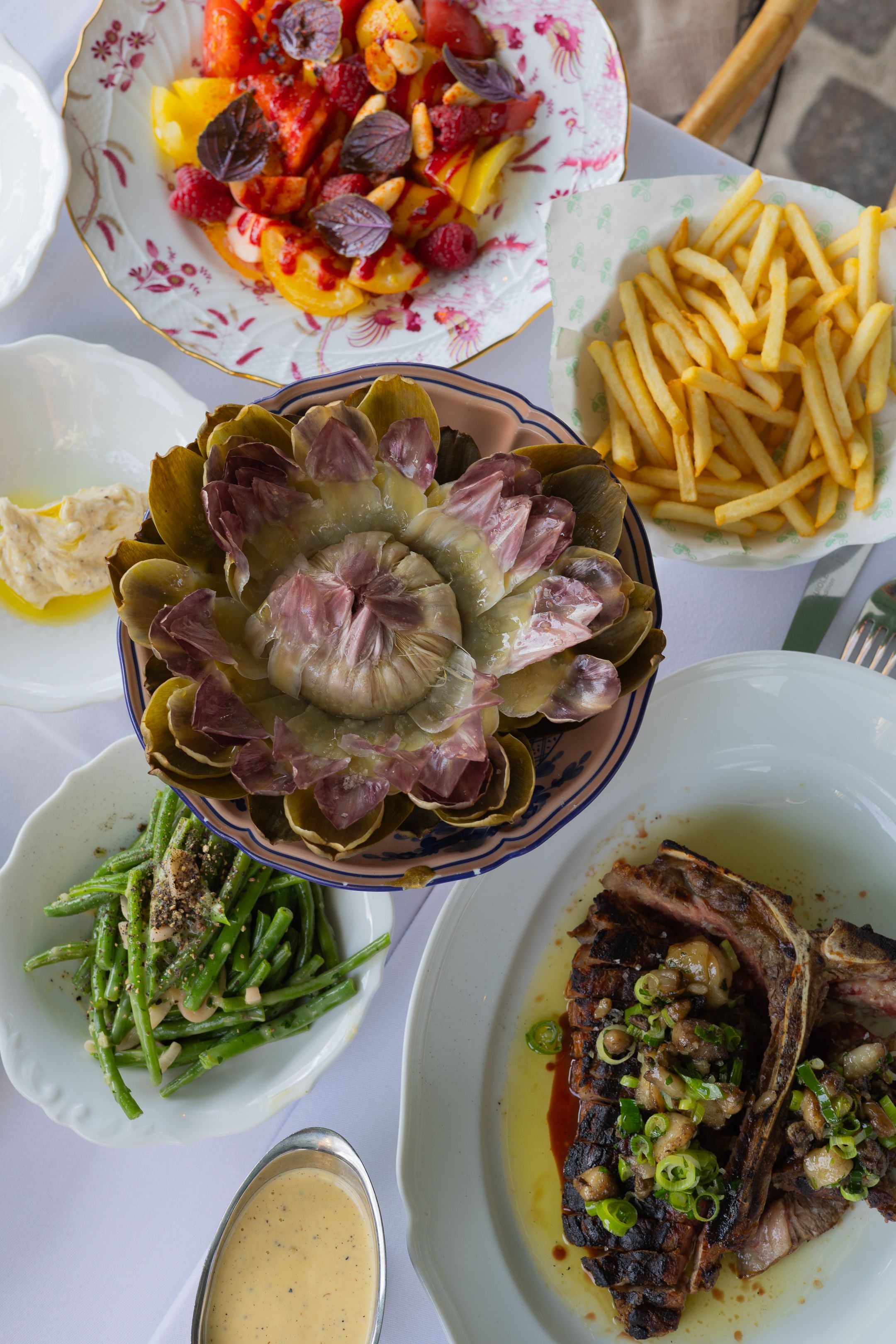 Elegant dinner spread featuring grilled artichoke, French fries, green beans, and meat dish with sauce on white plates.