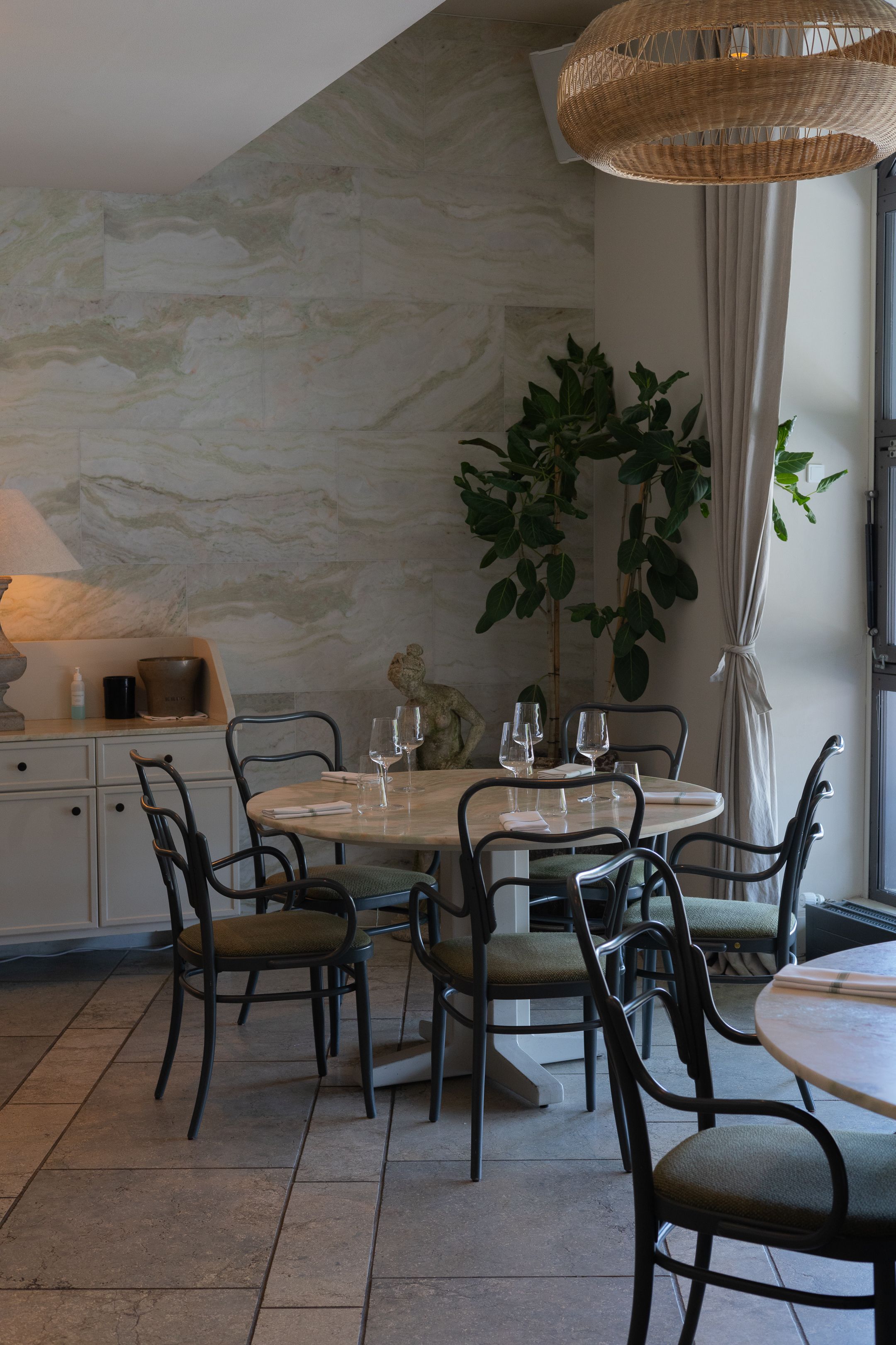 Modern dining area with round wooden table, black bentwood chairs, potted plant, and woven pendant light against marble wall.