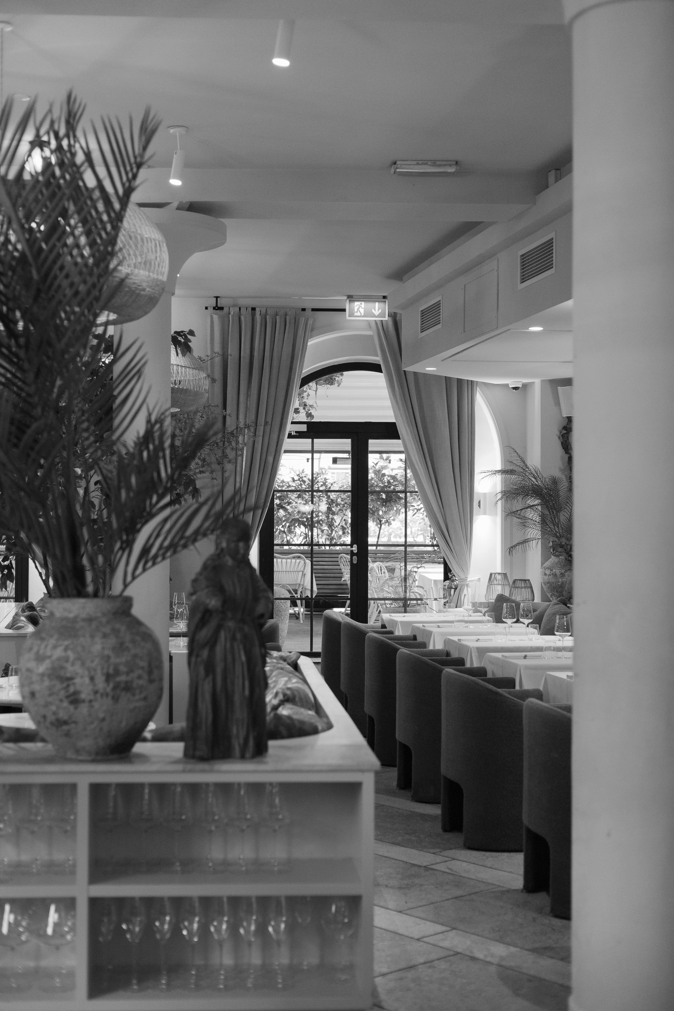 Black and white interior of an upscale restaurant with palm plants and booth seating.