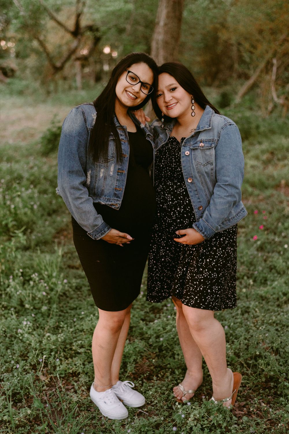 A candid outdoor photo of two people in denim jackets and dresses standing together in nature.