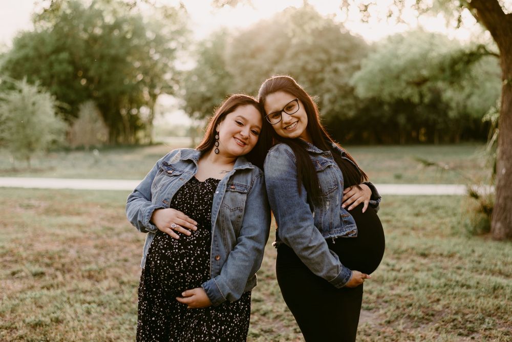 Two people in denim jackets lean on each other while posing in a park setting.