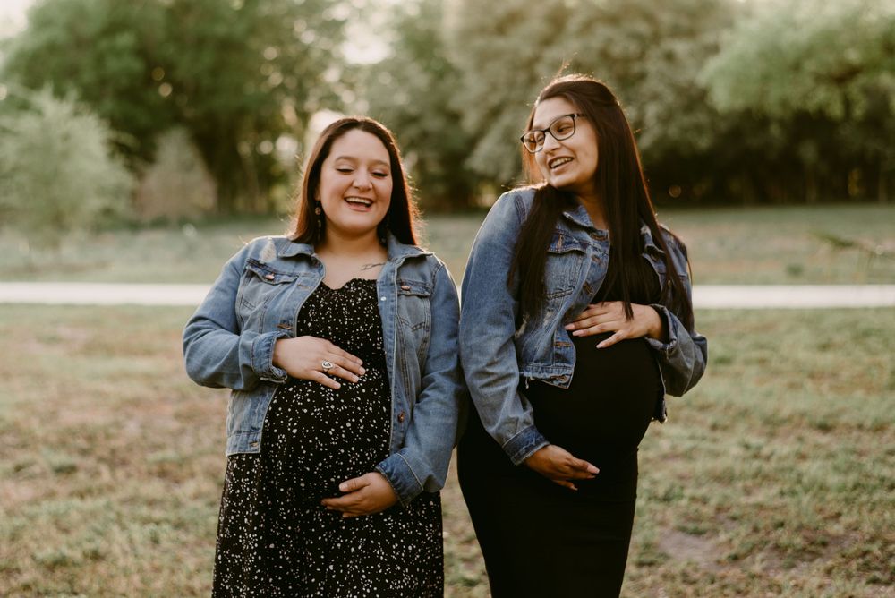 Two people in denim jackets share joyful expressions while touching their bellies outdoors.