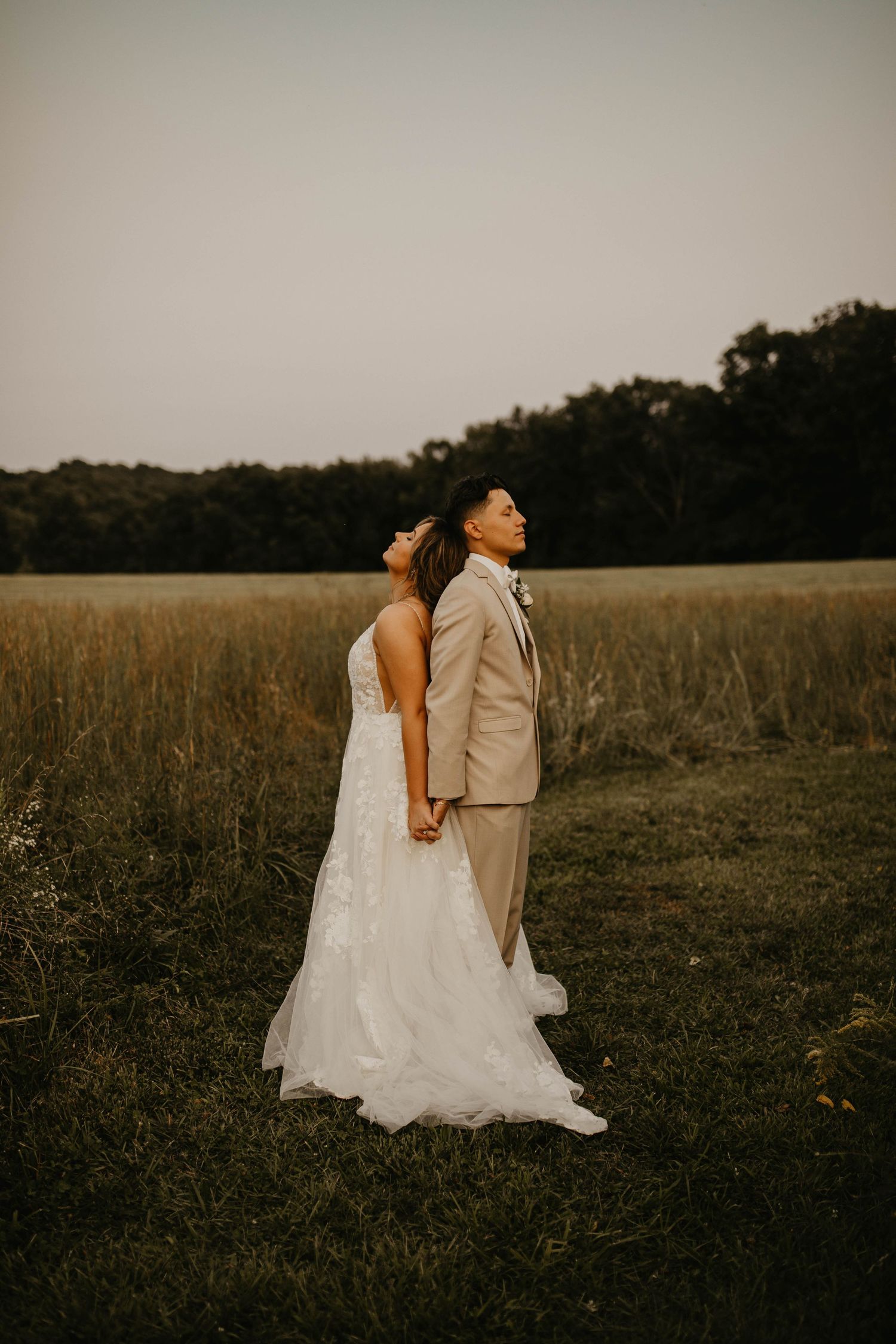 Newlywed couple standing back-to-back holding hands in golden field at sunset.