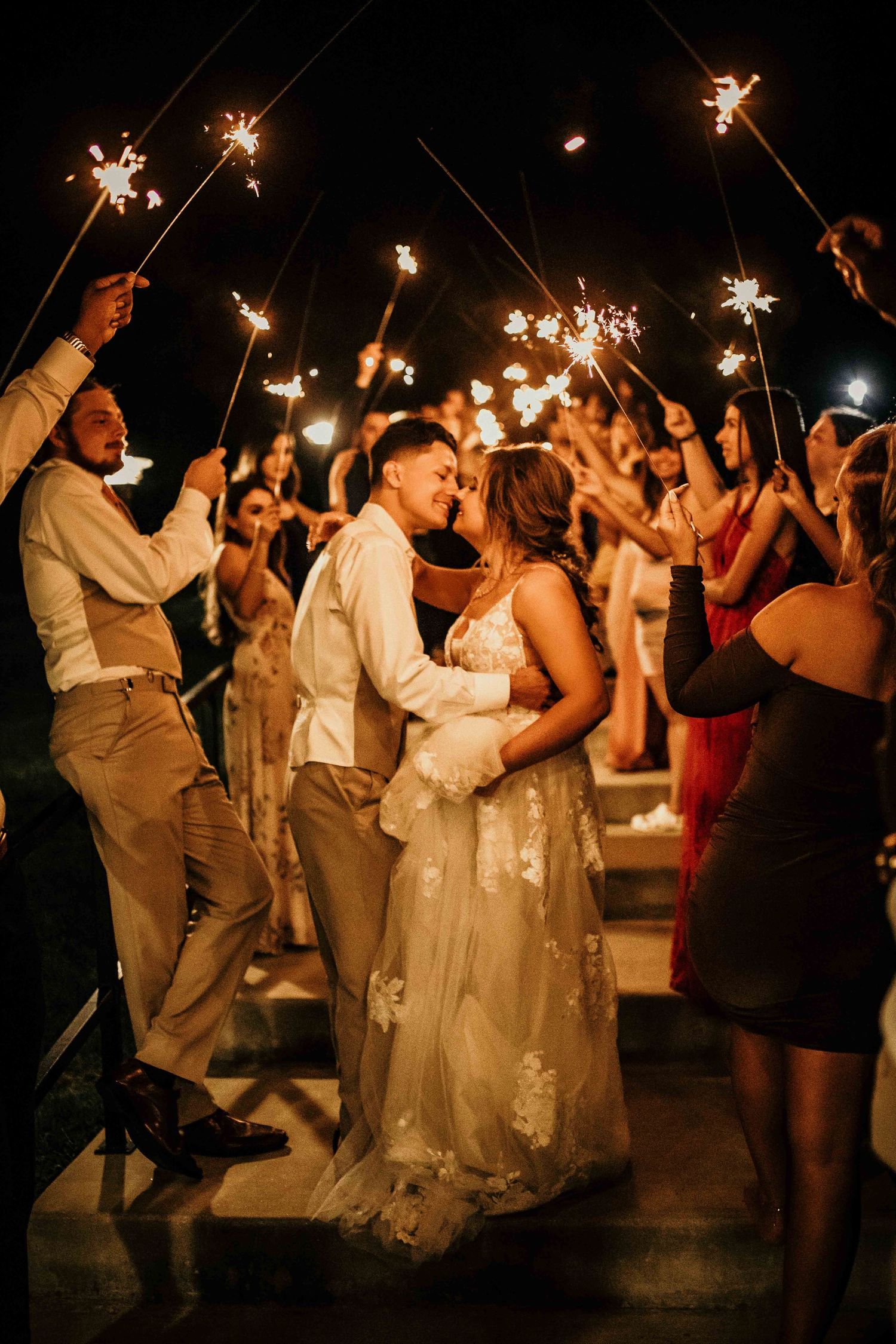 Newlyweds kissing during sparkler exit surrounded by cheering wedding guests at night celebration.