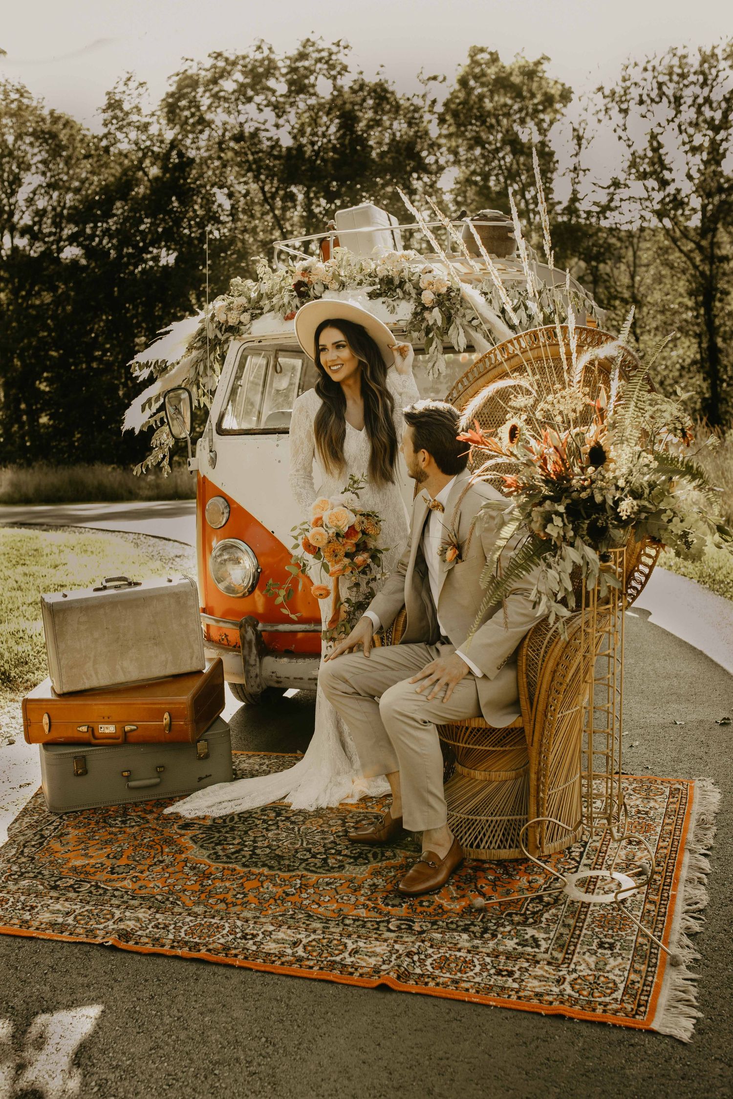 Boho bride and groom posing on vintage rug before orange VW bus with floral arrangements and luggage.