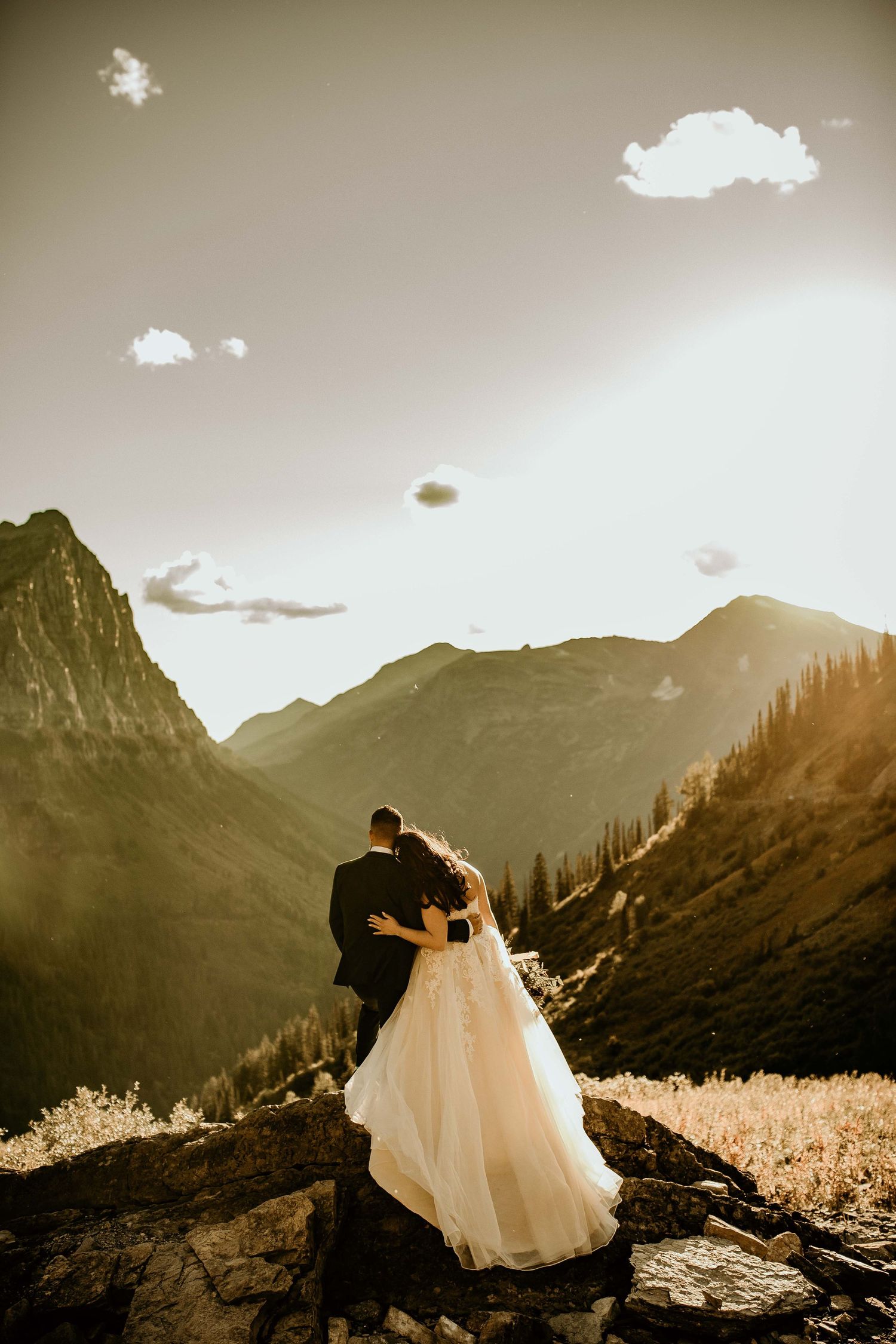 Wedding couple embracing on rocky mountain summit at golden sunset with dramatic alpine valley view.