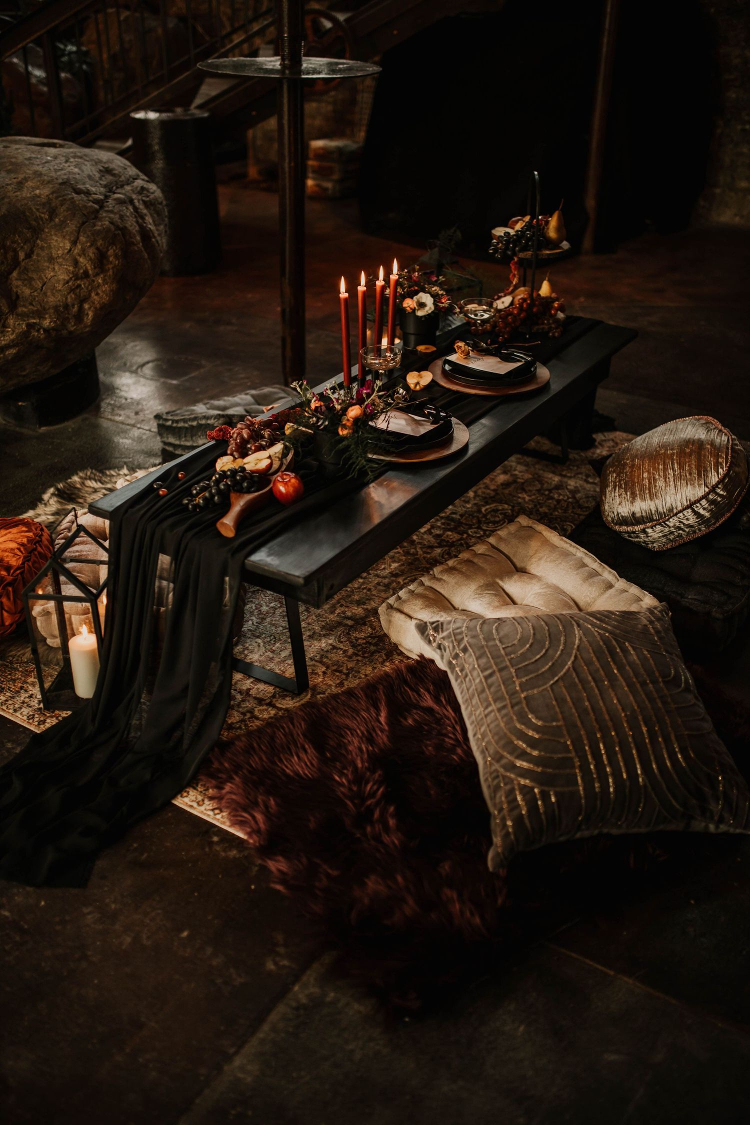 Dark moody wedding tablescape with red candles, floral centerpiece, fur rug, and velvet pillows.