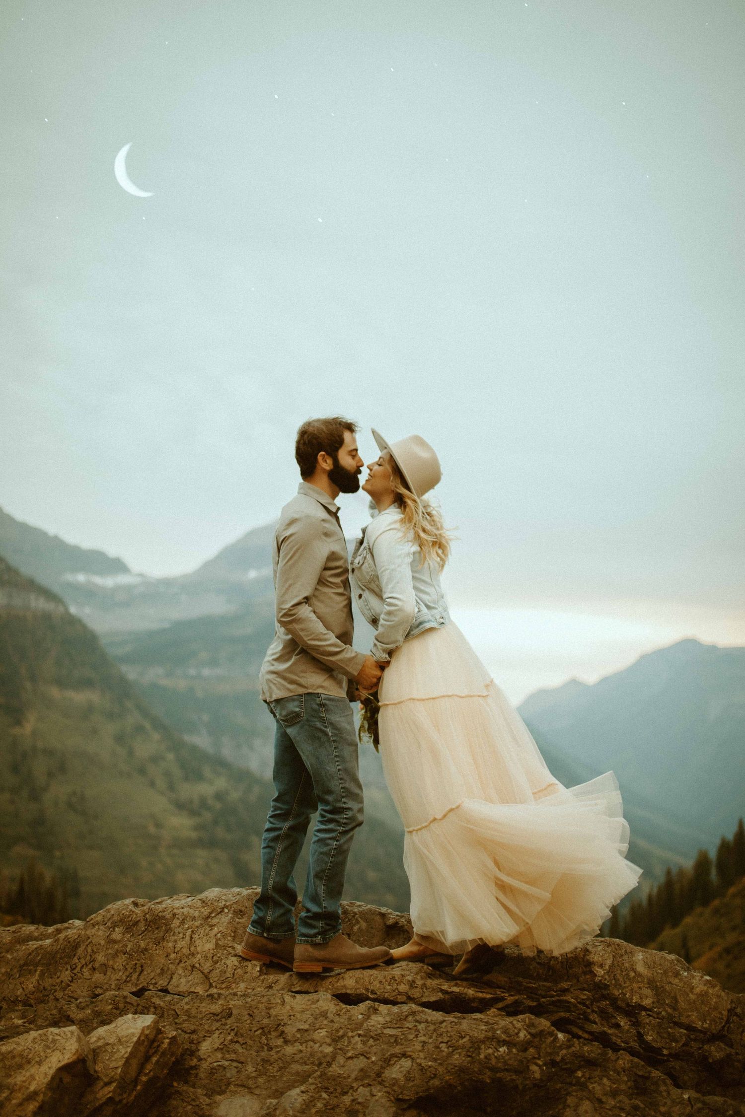 Couple kissing on mountain summit with crescent moon and sweeping valley views at dusk.