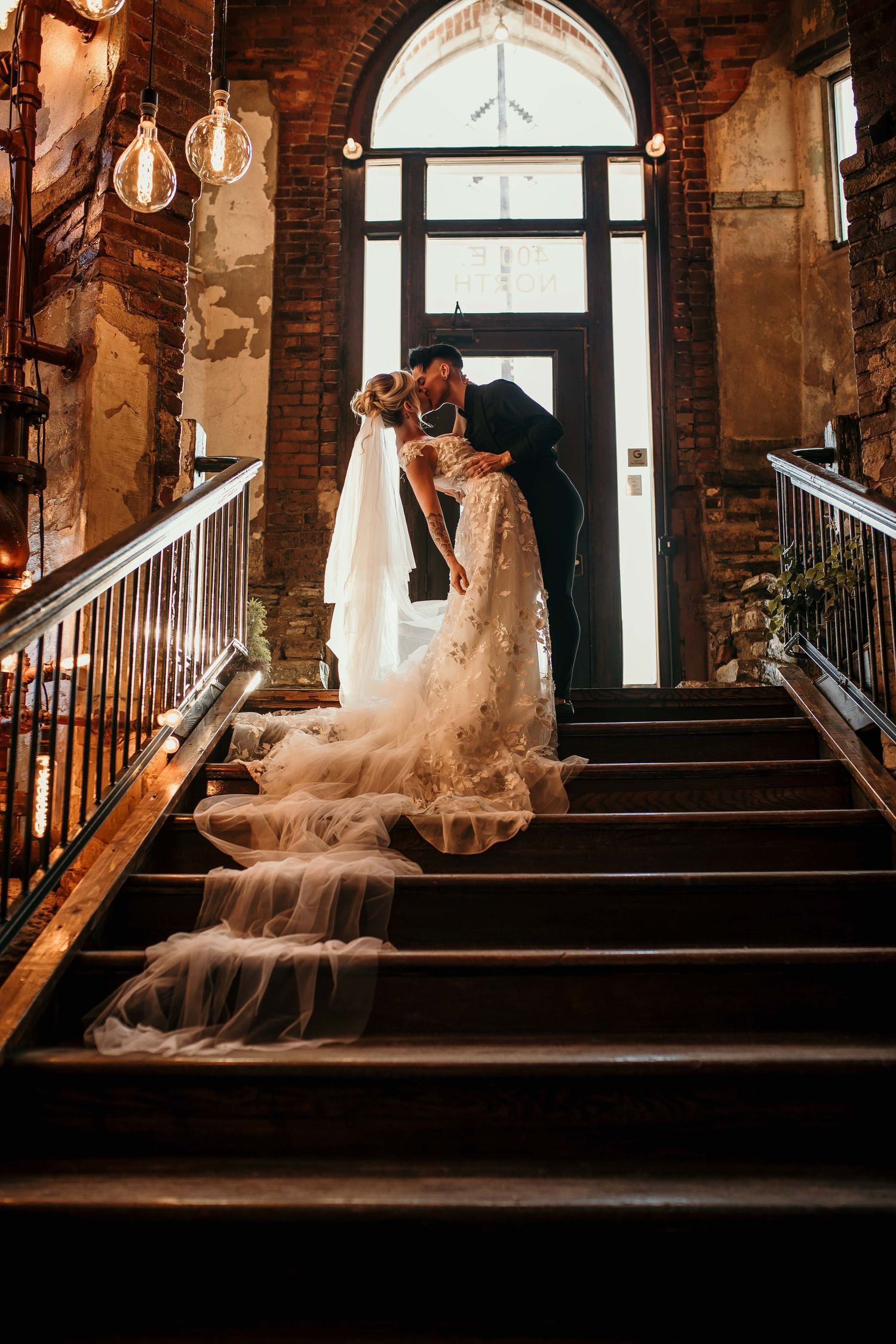Bride and groom sharing romantic kiss on grand staircase in rustic brick venue with flowing veil.