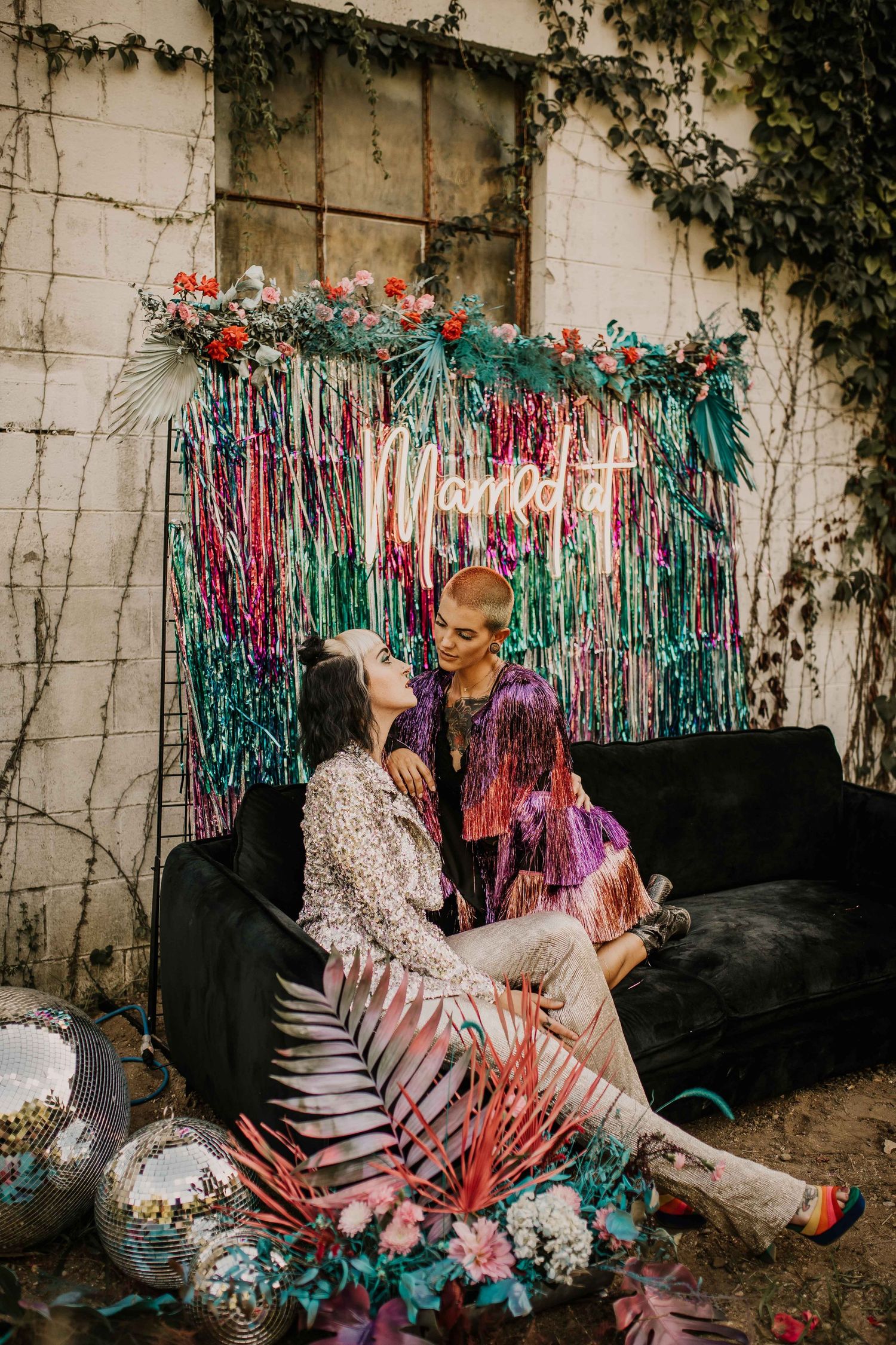 Two brides sit on a black velvet couch surrounded by colorful fringe backdrop and tropical florals at a bohemian wedding.