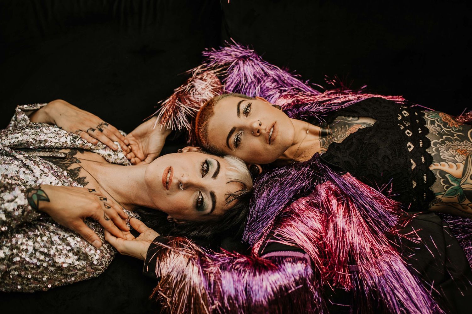 Three people lying together wearing colorful sequined and feathered outfits against black background.