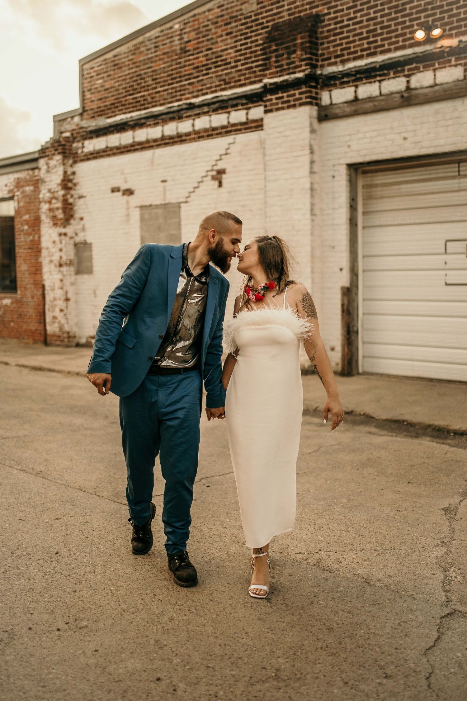 Tattooed bride in white midi dress and groom in blue suit sharing a kiss on urban street.