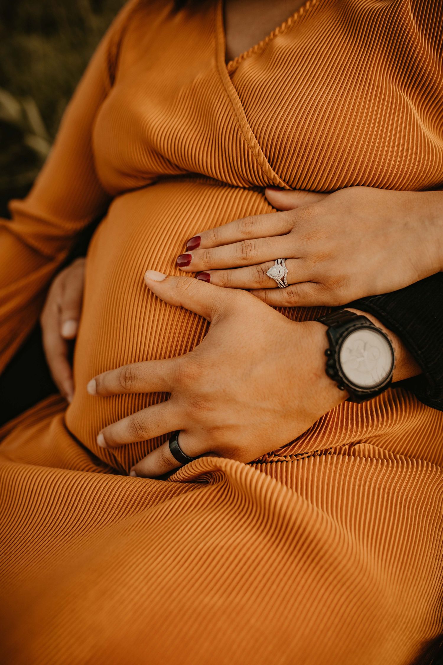 Close-up of pregnant woman in orange dress with couple's hands resting lovingly on baby bump.
