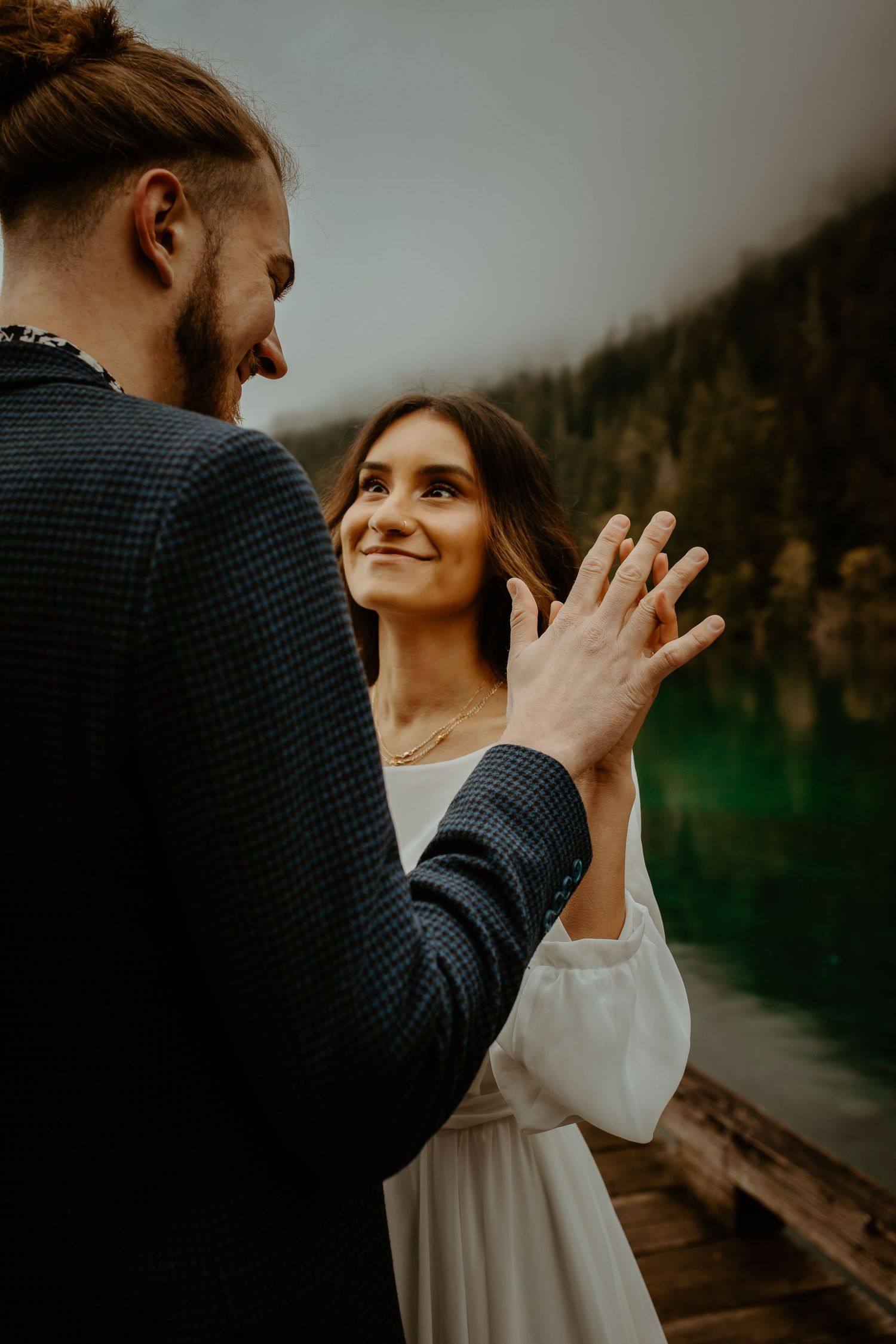 Bride smiling warmly as groom gently holds her hand on a lakeside dock.