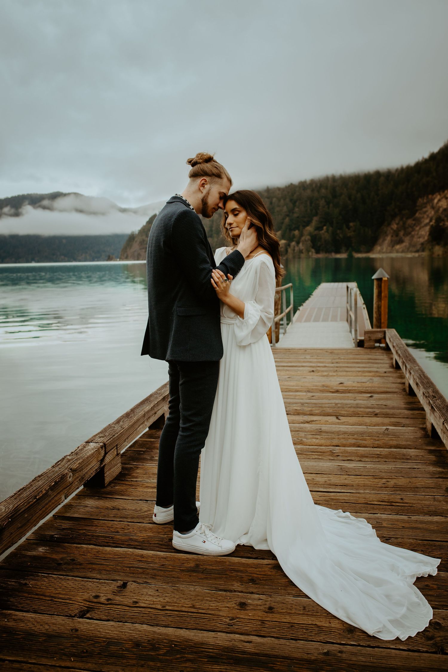 Bride and groom sharing a romantic moment on a wooden dock overlooking a turquoise mountain lake.