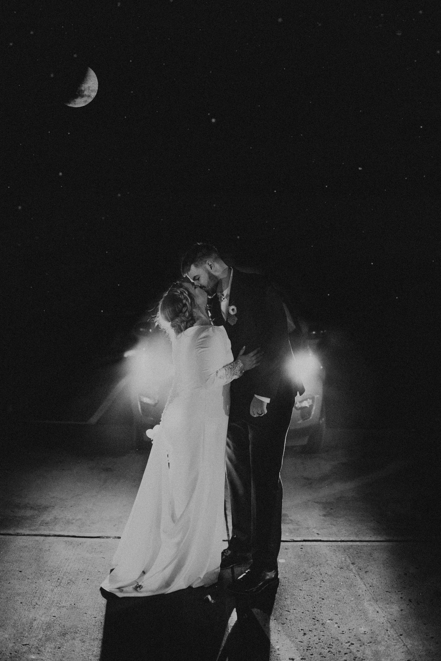 Black and white photo of newlyweds kissing under crescent moon and starry night sky with car lights.