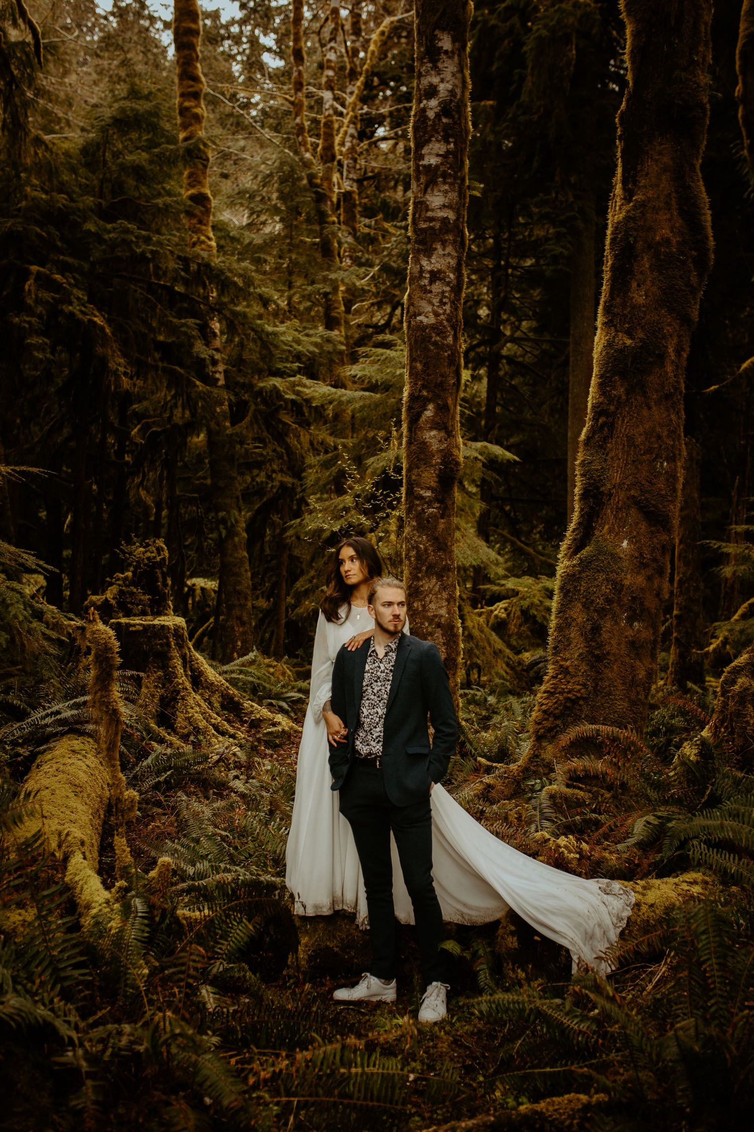 Bride and groom standing together in a moss-covered forest with a flowing white wedding dress.