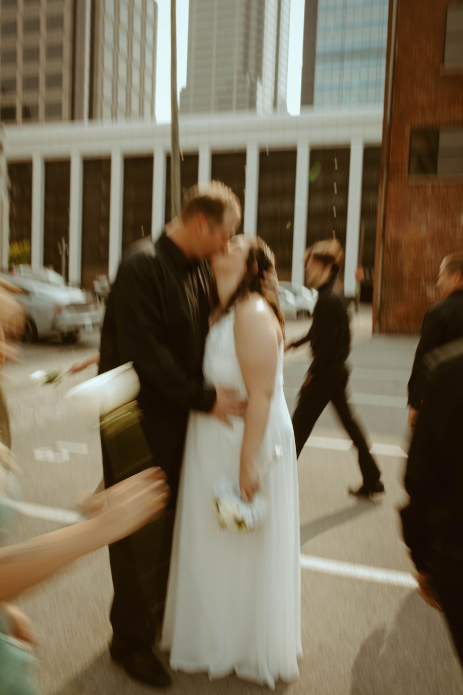 Bride and groom sharing a kiss on a busy city street in motion-blurred photo.